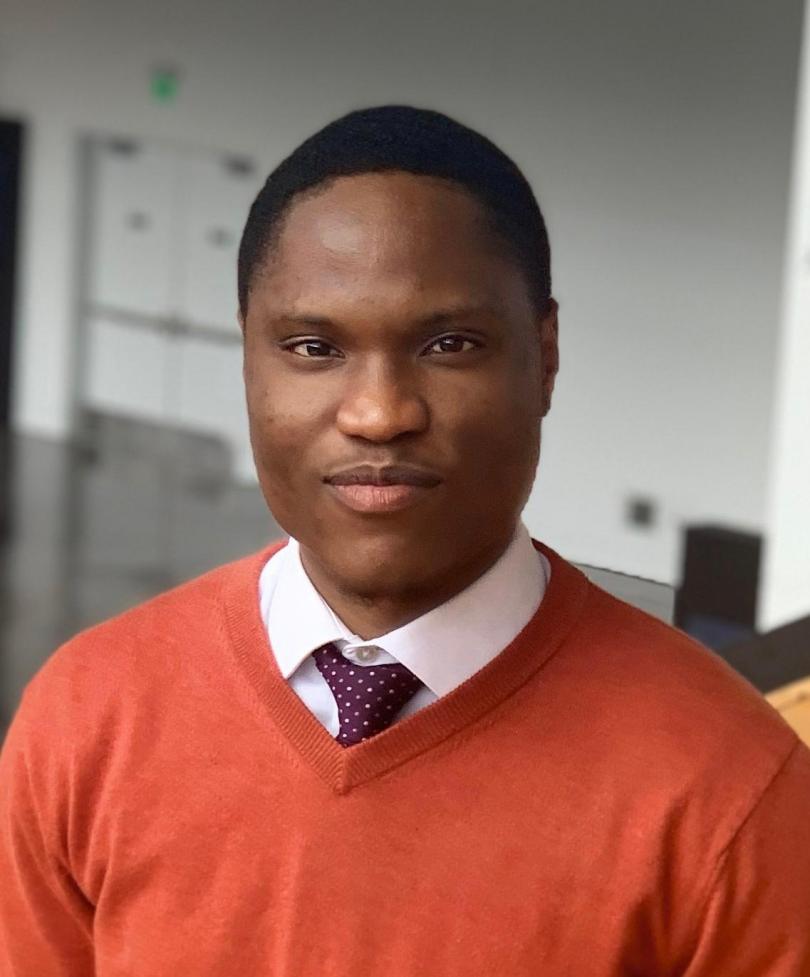 Here is short, compliant alt text:  **Alt text:** A man wearing an red sweater over a white shirt and polka-dot tie looks toward the camera in an indoor setting.