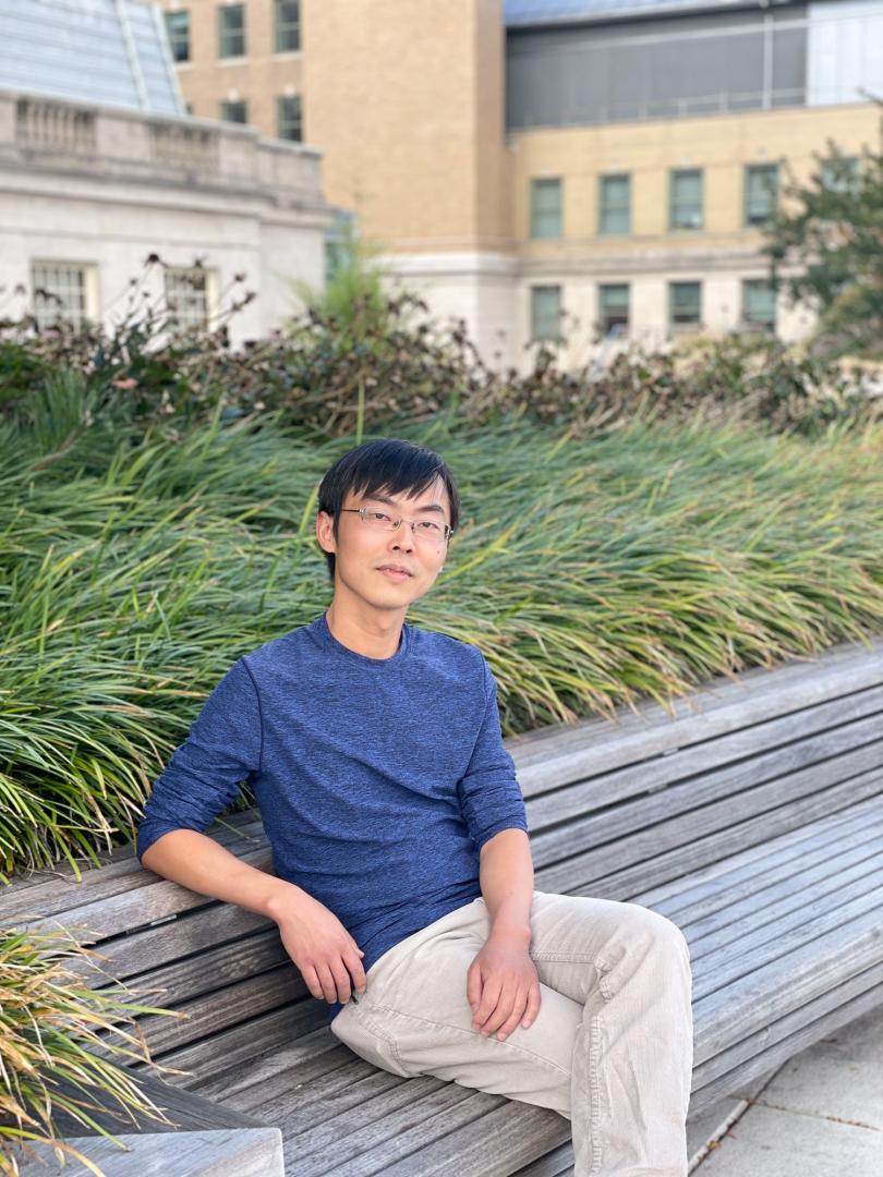 Photo of man with black hair and glasses wearing a blue long sleeved shirt and khaki pants sitting outside on a bench surrounded by greenery.