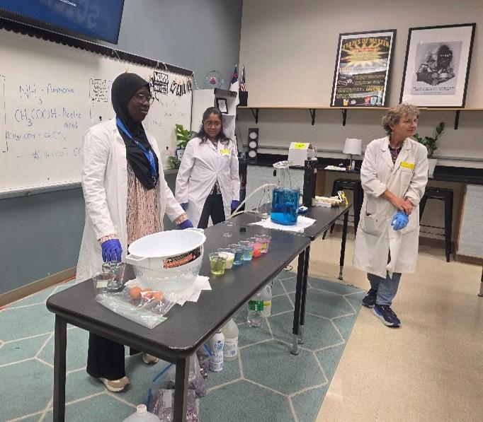 Three women in white lab coats and blue gloves conduct a chemistry demonstration at a table with colorful liquid-filled beakers and other supplies. A whiteboard with chemical equations is visible in the background.